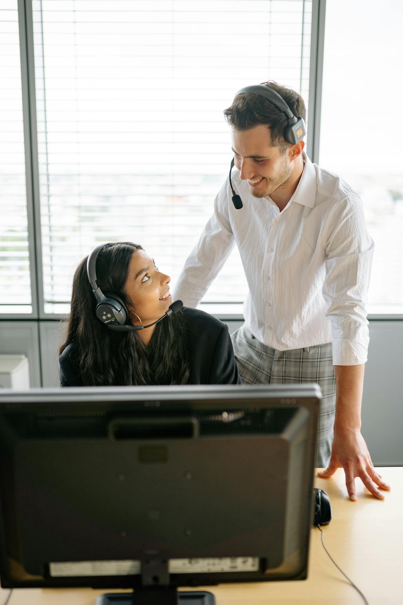 Two colleagues in headsets collaborating at a desk in a bright office setting.