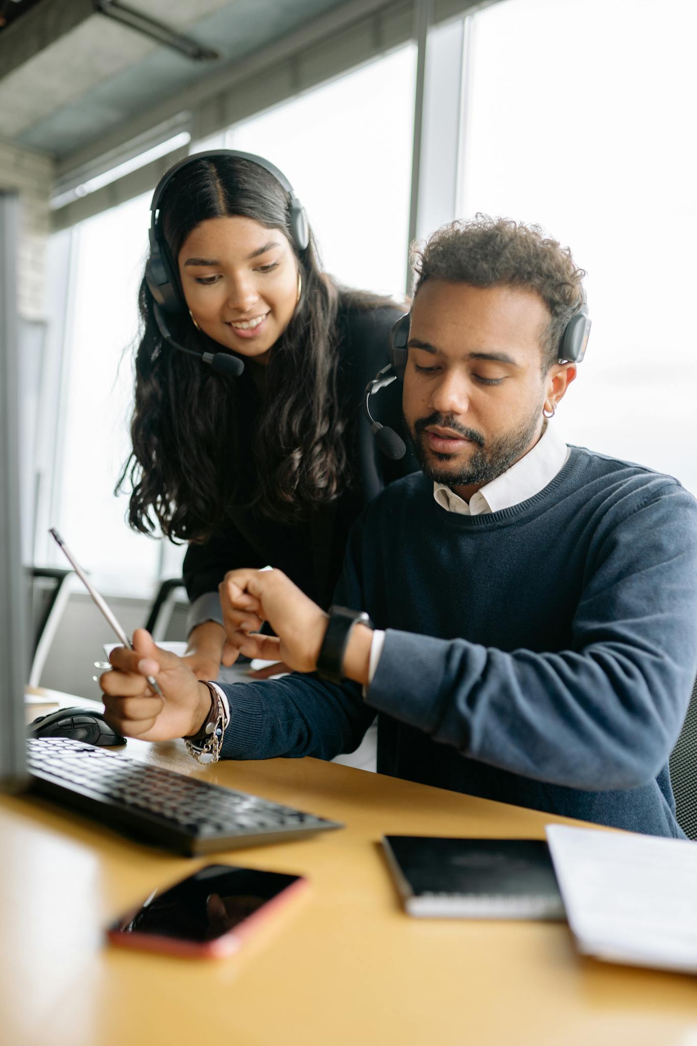 Two office workers wearing headsets collaborate at a desk, focusing on teamwork and communication.